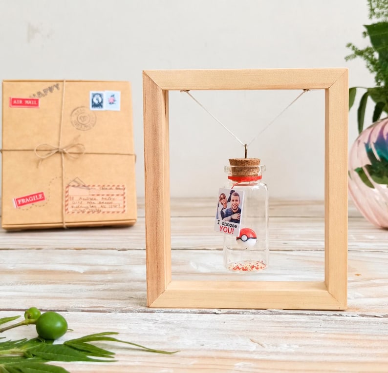 May include: A small glass bottle with a cork stopper hangs inside a light-colored wooden frame. Inside the bottle is a tiny red and white ball and a small rectangular tag featuring a photograph of two people and the text I choose YOU!