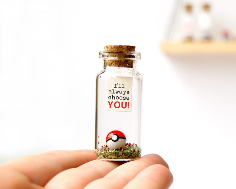 May include: A small glass jar with a cork lid filled with green and red glitter and a red and white Pokeball. A white paper note attached to the jar reads "I'll always choose YOU!"