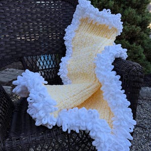 May include: A soft, hand-knitted blanket featuring a pale yellow center with a thick, textured white border made of looped yarn. The blanket has a plush, chunky appearance with a distinct contrast between the yellow body and the white ruffled edge.