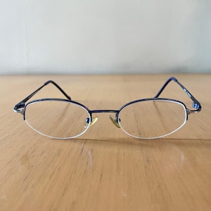 Puede incluir: Gafas de forma ovalada con montura azul oscuro y lentes transparentes. Las gafas tienen un diseño fino y delicado con un puente nasal dorado. Las patillas son negras y anguladas, y las lentes están delineadas en blanco.
