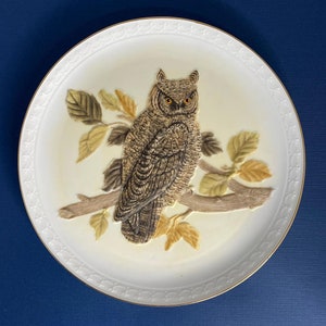 Puede incluir: Un plato de cerámica blanco con un borde dorado presenta una imagen en relieve 3D de un búho posado en una rama con hojas. El búho tiene plumas marrones y grises y ojos amarillos.