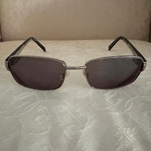 Peut inclure: Lunettes de soleil rectangulaires avec une monture argentée et des verres foncés. Les lunettes de soleil ont un design classique avec une monture fine et un pont sur le nez. Les branches sont noires.