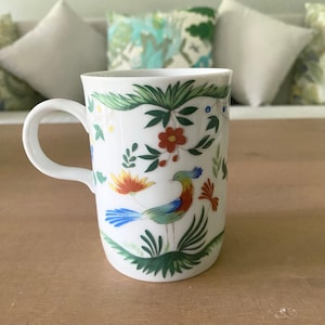 May include: White ceramic mug with a colorful floral and bird design. The design features a blue, green, yellow, and red bird with a long tail, surrounded by green leaves and red, yellow, and orange flowers.