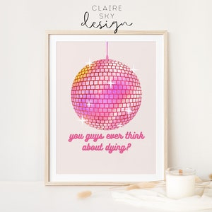 Puede incluir: Impresi&oacute;n enmarcada con una bola de discoteca rosa y amarilla y el texto "you guys ever think about dying?" en rosa. La impresi&oacute;n tiene un fondo rosa claro y est&aacute; firmada "Claire Sky Design". Una vela est&aacute; sobre la mesa.