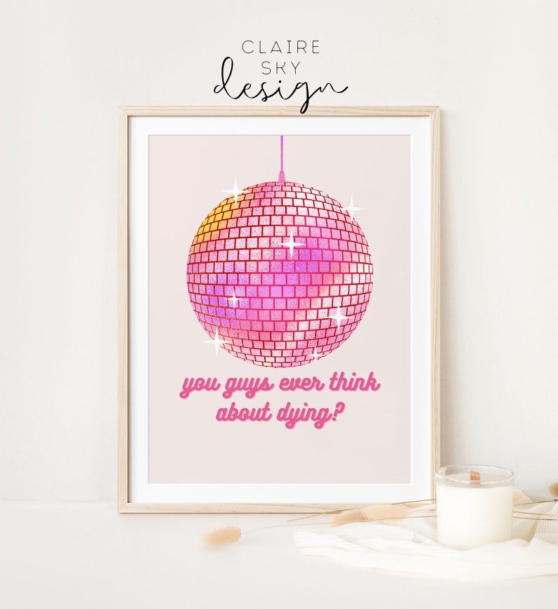 Puede incluir: Impresi&oacute;n enmarcada con una bola de discoteca rosa y amarilla y el texto "you guys ever think about dying?" en rosa. La impresi&oacute;n tiene un fondo rosa claro y est&aacute; firmada "Claire Sky Design". Una vela est&aacute; sobre la mesa.