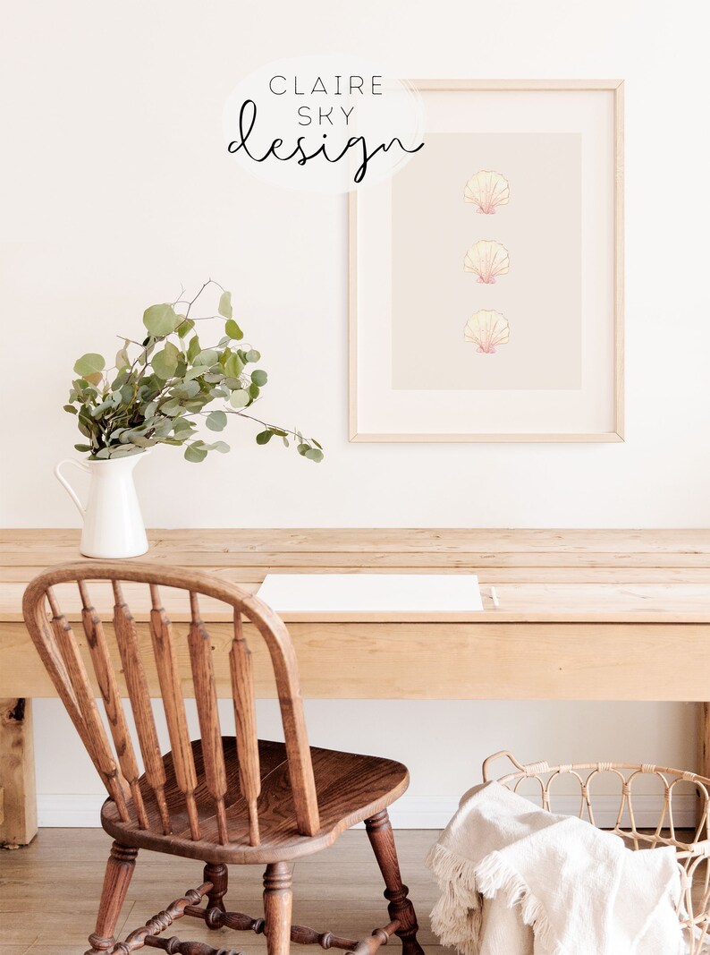 May include: A framed print with three pink and white seashells on a light gray background. The frame is light wood and the print is hanging on a white wall. There is a wooden table with a white pitcher and greenery in front of the print. A wooden chair and a white blanket are on the table.