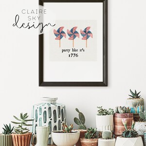 Op de afbeelding: Een ingelijst afdruk met een witte achtergrond en drie rood-wit-blauwe windmolens met de tekst "party like it's 1776". De afdruk is omgeven door een zwart frame en wordt weergegeven op een witte muur met verschillende potplanten ervoor.