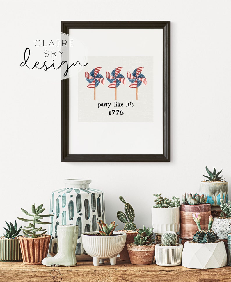 Op de afbeelding: Een ingelijst afdruk met een witte achtergrond en drie rood-wit-blauwe windmolens met de tekst "party like it's 1776". De afdruk is omgeven door een zwart frame en wordt weergegeven op een witte muur met verschillende potplanten ervoor.