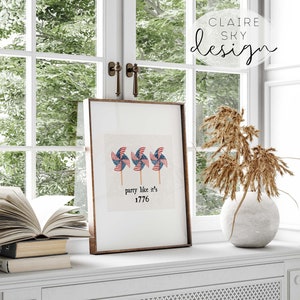 Op de afbeelding: Een ingelijst print met een witte achtergrond en drie rood, wit en blauwe windmolens. De tekst "party like it's 1776" staat onder de windmolens.