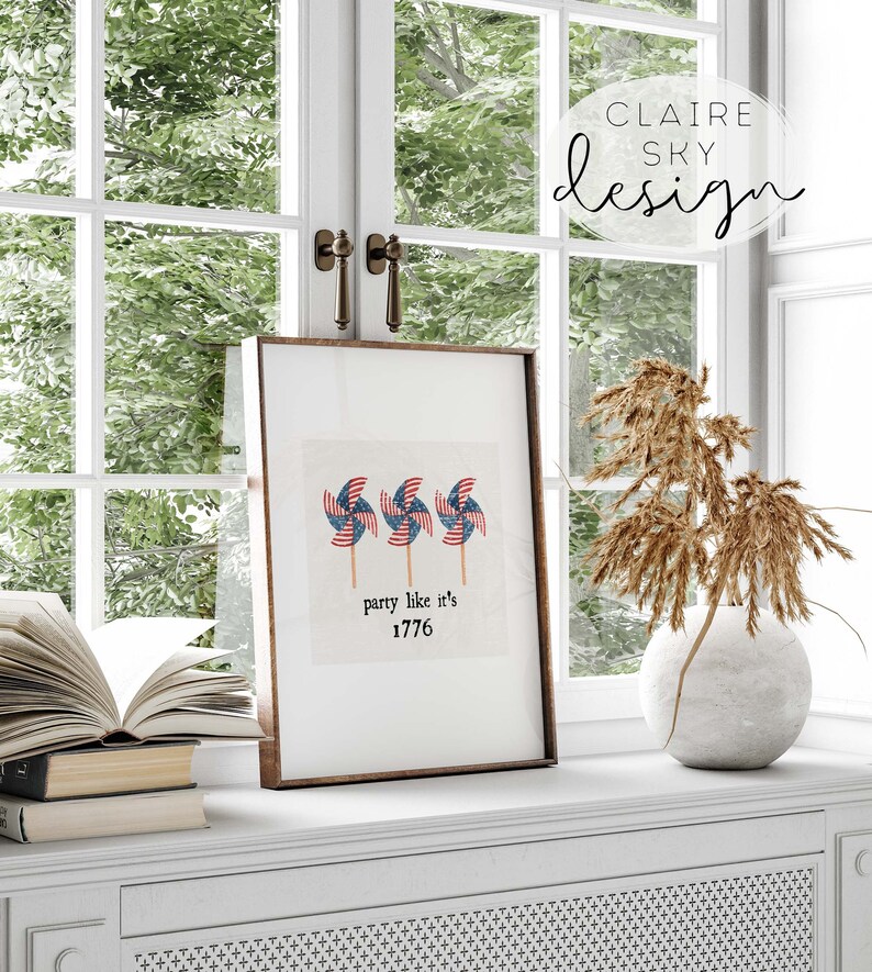 Op de afbeelding: Een ingelijst print met een witte achtergrond en drie rood, wit en blauwe windmolens. De tekst "party like it's 1776" staat onder de windmolens.