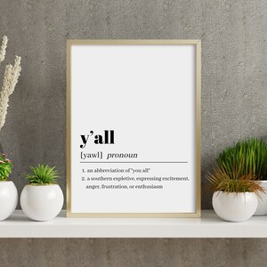 May include: A framed print with a white background and black text. The text defines the word "y'all" as a pronoun, an abbreviation of "you all", and a southern expletive expressing excitement, anger, frustration, or enthusiasm.