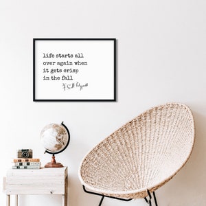 May include: Black and white framed print with a quote by F. Scott Fitzgerald that reads "life starts all over again when it gets crisp in the fall". The print is hanging on a white wall above a wicker chair and a globe.