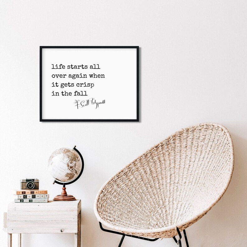 May include: Black and white framed print with a quote by F. Scott Fitzgerald that reads "life starts all over again when it gets crisp in the fall". The print is hanging on a white wall above a wicker chair and a globe.