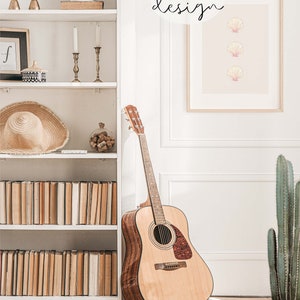 May include: A white bookshelf with books, a straw hat, a vase with dried flowers, and two candle holders. A brown acoustic guitar is leaning against the bookshelf. A framed print with three pink seashells on a light pink background is hanging on the wall. The print has the text 'CLAIRE SKY design' in a circle above the seashells. A cactus plant in a brown pot is on the floor next to the guitar.