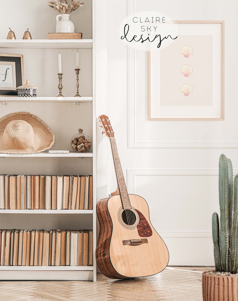 May include: A white bookshelf with books, a straw hat, a vase with dried flowers, and two candle holders. A brown acoustic guitar is leaning against the bookshelf. A framed print with three pink seashells on a light pink background is hanging on the wall. The print has the text 'CLAIRE SKY design' in a circle above the seashells. A cactus plant in a brown pot is on the floor next to the guitar.
