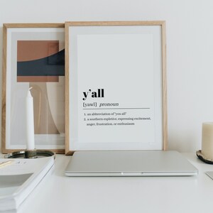 May include: A framed print with the word "y'all" defined as a pronoun, with the definition "an abbreviation of 'you all'" and "a southern expletive, expressing excitement, anger, frustration, or enthusiasm". The print is on a white desk with a laptop, a candle, and a stack of books.