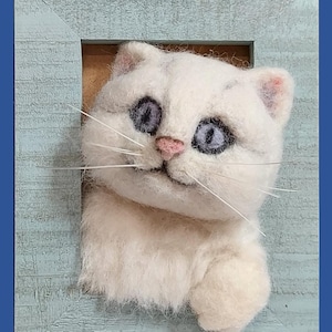 Può includere: Scultura di un gatto bianco con occhi blu e naso rosa, che emerge da una cornice di legno azzurro. Il gatto ha pelo soffice e baffi. L'opera d'arte è ambientata su uno sfondo blu, creando un effetto affascinante e stravagante.