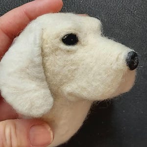 May include: A handcrafted, off-white felt dog head with a black nose and eyes. The dog's head is held in a hand, showcasing the detailed craftsmanship of the felted sculpture. The dog's ears are floppy and the fur is textured.