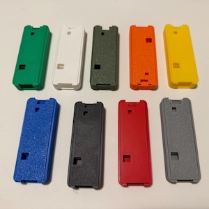 Puede incluir: Una colección de nueve carcasas rectangulares de plástico en varios colores, incluyendo verde, blanco, oliva, naranja, amarillo, azul, negro, rojo y gris. Cada carcasa tiene un diseño similar con un recorte rectangular y una pequeña abertura cuadrada.