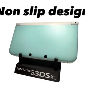 Peut inclure: Console de jeu portable Nintendo 3DS XL turquoise sur un support noir. Le support porte l'inscription "NINTENDO 3DS XL" en blanc. Le texte "Non slip design" est au-dessus.