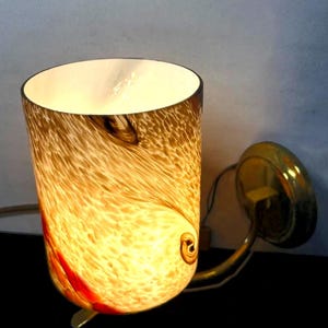 May include: A wall-mounted sconce with a cylindrical, speckled glass shade in shades of brown, amber, and red. The interior is white, and the fixture has a brass-colored metal arm and mounting plate. The light is on, illuminating the glass.