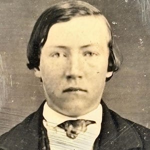 May include: A vintage portrait of a person in a dark suit with a white collared shirt and a patterned tie. The image is a close-up shot, with the person looking directly at the camera. The photograph has a sepia tone and shows signs of age.