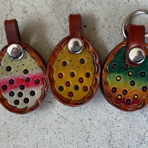 May include: Three leather keychains with different colored patterns. Each keychain features a round, brown leather tag with a silver metal ring. The patterns include a white and pink striped design, a yellow and red polka dot design, and a green, yellow, and red gradient design.