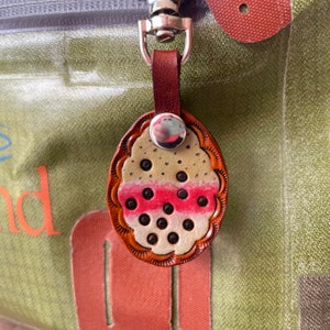Puede incluir: Un llavero de cuero con un dise&ntilde;o estampado en rosa y blanco. El llavero tiene forma de una peque&ntilde;a fruta redonda con un anillo plateado en la parte superior.
