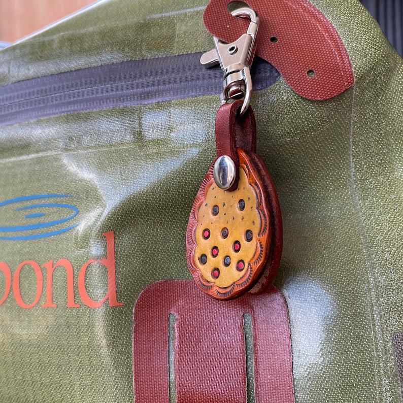 Puede incluir: Un llavero de cuero marr&oacute;n con un charm decorativo de patr&oacute;n amarillo y rojo. El charm tiene forma de l&aacute;grima y un anillo de metal plateado.