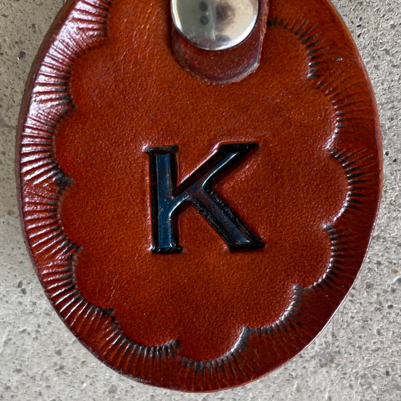 Puede incluir: Un llavero de cuero marr&oacute;n con una letra K negra en relieve en la parte delantera. El llavero tiene un borde festoneado decorativo.