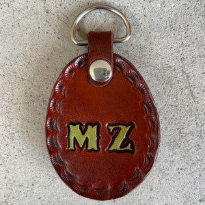 Puede incluir: Llavero de cuero marr&oacute;n con un anillo plateado y las iniciales "MZ" en letras verdes.