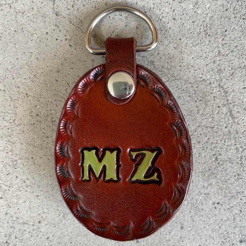 Puede incluir: Llavero de cuero marr&oacute;n con un anillo plateado y las iniciales "MZ" en letras verdes.