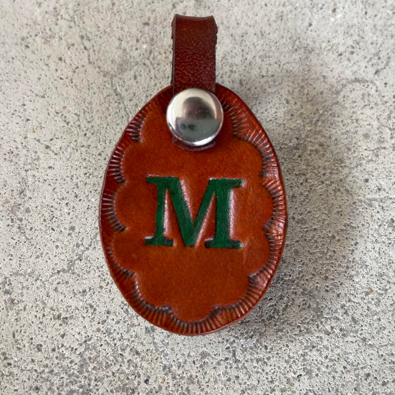 Puede incluir: Un llavero de cuero marr&oacute;n con un anillo de metal plateado y una letra M verde en la parte delantera.
