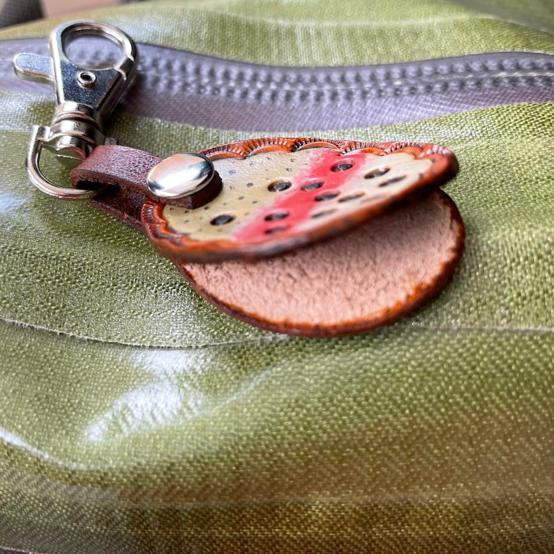 Puede incluir: Un llavero de cuero con un dise&ntilde;o detallado de una trucha. El llavero es marr&oacute;n con un patr&oacute;n marr&oacute;n claro y rojo. El llavero tiene un anillo de metal plateado y un cierre de metal plateado.