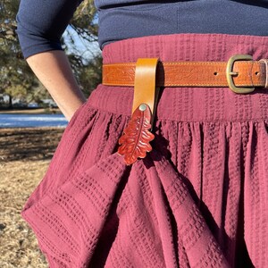 May include: A maroon skirt with a brown leather belt and a leather leaf-shaped belt buckle. The belt buckle is a reddish-brown color.