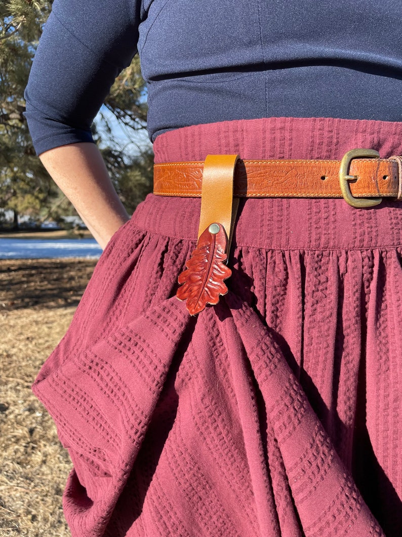 May include: A maroon skirt with a brown leather belt and a leather leaf-shaped belt buckle. The belt buckle is a reddish-brown color.