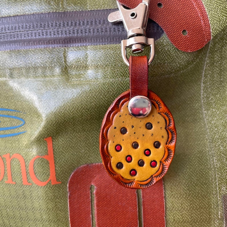 Puede incluir: Un llavero de cuero marr&oacute;n con un dije redondo de patr&oacute;n amarillo y rojo. El dije tiene un borde festoneado y un anillo plateado.