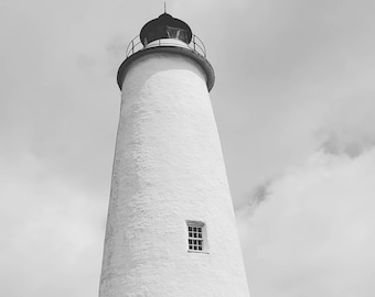 May include: A tall, white lighthouse with a black top and a circular railing. The lighthouse has two small windows and is set against a cloudy sky. The base of the lighthouse is surrounded by trees.