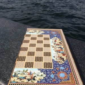 May include: A rectangular game board featuring a central grid of alternating light and dark patterned squares. The border is decorated with intricate blue, gold, and white floral motifs and detailed miniature paintings of figures on horseback.