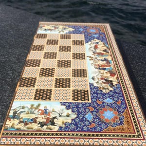 Könnte beinhalten: Ein rechteckiges Backgammon-Spielbrett mit einem zentralen Raster aus abwechselnd schwarzen und beigen gemusterten Quadraten. Das Brett ist mit aufwendigen blauen Blumenbordüren und handgemalten Szenen von Reitern in einer Landschaft verziert.