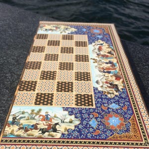 May include: A rectangular wooden game board featuring a central grid of alternating black and beige squares with intricate floral patterns. The border is decorated with blue, gold, and white motifs, including detailed miniature paintings of figures.