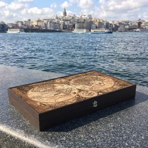 Könnte beinhalten: Eine rechteckige, dunkelbraune Holzbox mit detailliertem Kartendesign auf dem Deckel. Die Box steht vor einer Stadtlandschaft und Wasser, unter einem strahlend blauen Himmel.