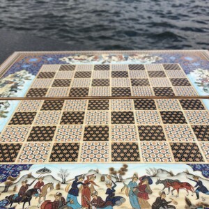 May include: A rectangular folding chessboard featuring a central grid of alternating dark brown and light beige squares with intricate geometric patterns. The border displays detailed miniature paintings of figures in traditional attire, horses, and landscapes.