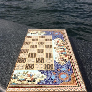 May include: A rectangular chessboard featuring a central grid of alternating light and dark squares with intricate geometric patterns. The border is decorated with colorful miniature paintings of figures on horseback and floral motifs in blue, gold, and red.