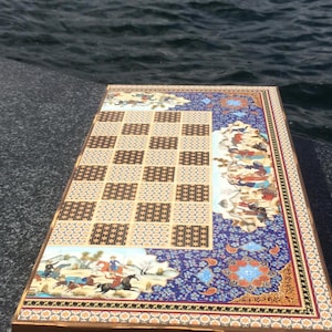 May include: A decorative wooden game board with a chessboard design and intricate Persian artwork. The board features a checkerboard pattern, detailed illustrations, and a blue floral border. The board is set against a backdrop of water.