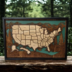 May include: A framed wooden map of the United States, featuring a detailed relief design. The map is in a dark wood frame, with the states and surrounding landscape carved in varying shades of brown and tan. The background is a teal color, and the map is set against a blurred forest backdrop.