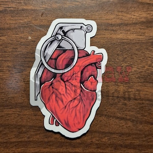 May include: A red anatomical heart with a gray grenade pin and handle. The heart is depicted as a grenade.