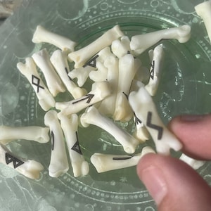 May include: A collection of white bone dice with black runes painted on them. The dice are in a green bowl with a decorative pattern.