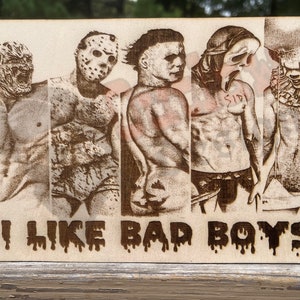 May include: A wooden sign with a brown background and a black and white image of five horror movie characters. The characters are wearing masks and are standing in a row. The text "I LIKE BAD BOYS" is written in black below the image.