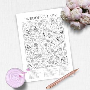 May include: A black and white "Wedding I Spy" game sheet with various wedding-themed illustrations and a list of items to find. The sheet is on a white surface, with a pink drink and a rose gold pen in the foreground.